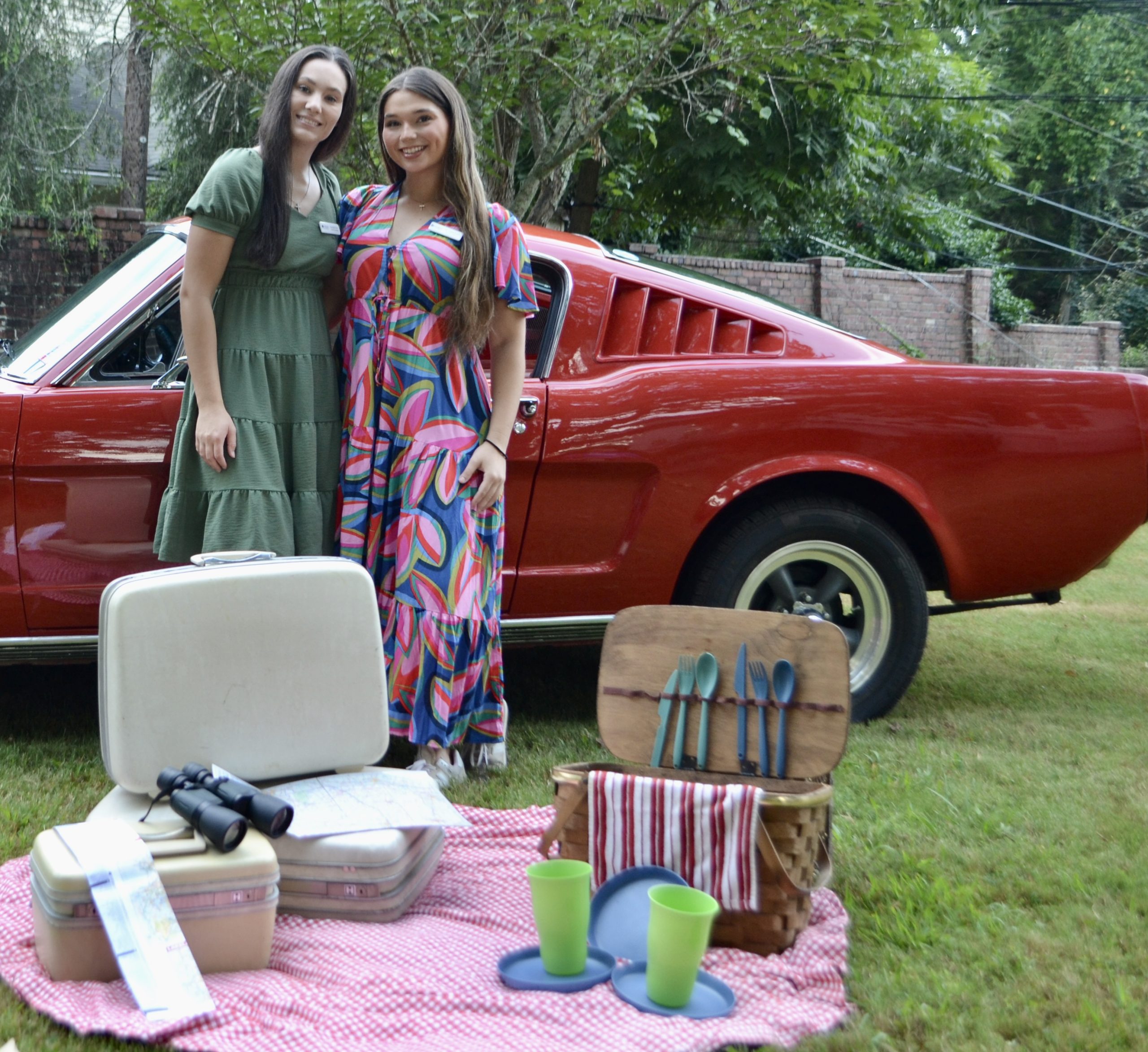 Two School of Education students stand smiling in front of a classic red car during a picnic setup, with vintage suitcases, a map, and a picnic basket laid out on a red-and-white blanket.